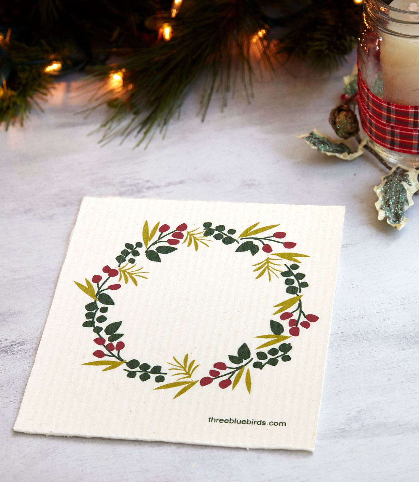 Winter Wreath Three Bluebirds Swedish Dishcloth next to green garland and a candle.