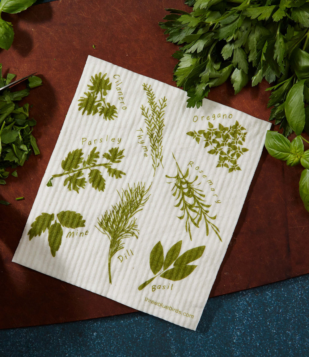 Herbs Swedish Dishcloth next to fresh cilantro, parsley, oregano, dill, basil, thyme, and mint.